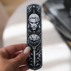 May include: A black and white bookmark featuring a detailed illustration of a stern-faced man with long white hair. The design includes a compass rose, a sword, and ornate floral elements, creating a gothic aesthetic.