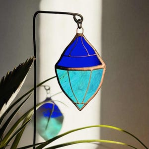 Può includere: Un acchiappasole in vetro colorato a forma di diamante, con una combinazione di blu scuro e chiaro, bordi color rame. È appeso a un gancio di metallo nero. Una pianta è visibile in primo piano.