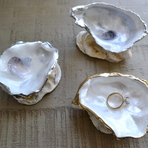 Op de afbeelding: Drie decoratieve oesterschelpen ringhouders. Twee schelpen hebben zilverkleurige randen en één heeft een goudkleurige rand. Eén schelp bevat een gouden ring met een diamant. De schelpen zijn wit.