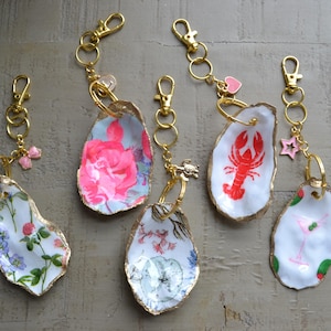 Pode incluir: Uma coleção de seis chaveiros decorativos de concha de ostra. Cada concha é pintada com um design único, incluindo padrões florais, uma lagosta vermelha e um coquetel rosa. As conchas têm bordas e ferragens douradas.