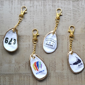 May include: Four keyrings crafted from oyster shells, each with gold-coloured fixings. The shells feature various designs: a number, a Boston logo, a colourful hot air balloon, and an island silhouette with the letters "ack".