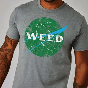 Può includere: T-shirt grigia con un design a cerchio verde con la scritta bianca "WEED", uno swoosh blu e un ovale bianco. Il design ricorda il logo della NASA.