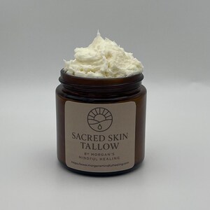 May include: A small, brown glass jar containing a white, whipped cream-like substance. The jar is labelled "SACRED SKIN TALLOW" and "BY MORGAN'S MINDFUL HEALING". This product is likely a skincare item.