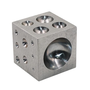 May include: A silver-colored metal dapping block cube featuring multiple hemispherical indentations of varying sizes on its top and side faces. The block has a brushed metallic finish and is designed with a large concave depression on the front.