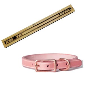 May include: A wooden die-cut tool with a series of small holes and a pink dog collar with a gold-colored buckle. The collar is made of a smooth, shiny material.