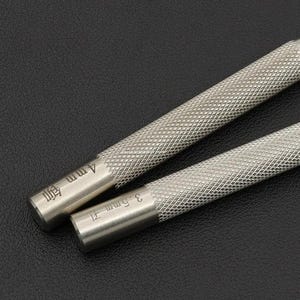 May include: Two silver metal tools with textured handles. One tool is labelled "4mm" and the other "3.5mm". The tools are angled diagonally across the frame, suggesting they are for crafting or precision work.