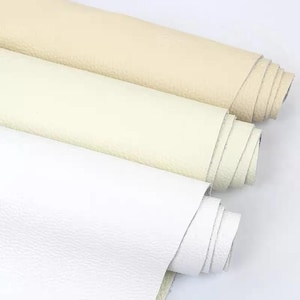 Puede incluir: Tres rollos de cuero sintético texturizado en crema, blanco roto y blanco. Los rollos están bien enrollados, revelando los bordes del material. La superficie del cuero tiene una textura sutil y granulada. Los colores son suaves y neutros.