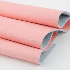 Puede incluir: Primer plano de varios rollos de tela de cuero sintético rosa. La tela tiene una superficie texturizada y un respaldo gris. Los rollos están en capas, mostrando las variaciones de textura y color. Ideal para proyectos de artesanía y tapicería.