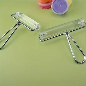May include: Two craft rollers with clear acrylic cylinders and silver metal handles. Each tool features a smooth, transparent barrel attached to a sturdy wire frame, designed for flattening materials during various creative projects.