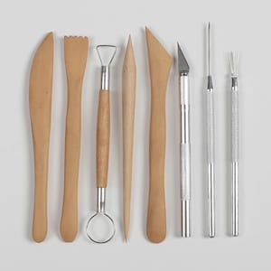 Könnte beinhalten: Ein Set aus sieben vertikal angeordneten Modellierwerkzeugen. Die Sammlung umfasst fünf hellbraune Holzinstrumente in verschiedenen Formen, darunter ein Messer, eine gezackte Kante, eine Schlaufe, eine Spitze und eine flache Klinge sowie zwei silberne Metallwerkzeuge.