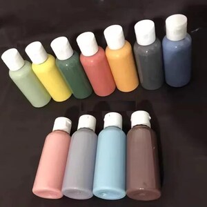 Puede incluir: Una colección de once botellas cilíndricas de plástico exprimibles llenas de varios colores de pintura líquida opaca. Cada botella está rematada con una tapa de rosca blanca. Los colores incluyen tonos de verde claro, amarillo, salvia, coral, naranja, gris, azul, rosa, lavanda, azul cielo y marrón.