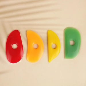 May include: Four kidney-shaped pottery ribs made of smooth plastic, each featuring a circular hole in the centre. The set includes one red, one orange, one yellow, and one green tool, all designed with curved edges for shaping and smoothing clay.