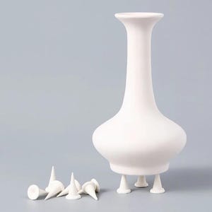 Puede incluir: Un jarrón de cerámica blanco mate con cuerpo bulboso y cuello alto y delgado. El jarrón se apoya sobre tres pequeños pies cónicos. Junto al jarrón hay varios pies cónicos blancos sueltos, idénticos en forma y tamaño a los de la base.