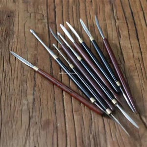 Könnte beinhalten: Ein Set aus sieben Modellierwerkzeugen mit langen, schlanken Holzgriffen in Dunkelbraun und Schwarz. Jedes Werkzeug hat eine Metallspitze, darunter spitze, abgerundete und flache Formen, die durch eine messingfarbene Metallhülse verbunden ist.