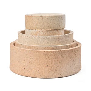 Peut inclure: Un ensemble de quatre anneaux circulaires en pierre poreuse de couleur beige clair, empilés par taille décroissante. Chaque anneau présente une surface rugueuse et texturée avec de petites taches sombres visibles, formant une structure en gradins.