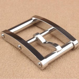 Puede incluir: Hebilla de cinturón plateada y brillante, con un marco rectangular y una barra central. La hebilla tiene bordes redondeados y está asegurada con pequeños remaches. El diseño es simple y moderno, adecuado para varios estilos de cinturón.