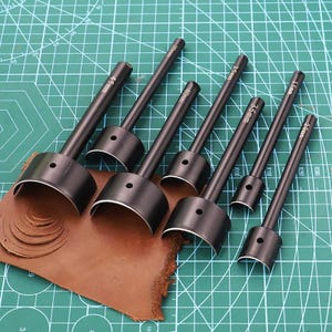 Könnte beinhalten: Ein Satz schwarzer Lederhandwerkzeuge in verschiedenen Größen, darunter 1,5 mm, 2,0 mm, 2,5 mm, 3,5 mm, 4,0 mm, 4,5 mm und 20 mm. Die Werkzeuge liegen auf einer grünen Schneidematte mit einem Stück braunem Leder. Diese Werkzeuge werden zum Schneiden von Leder verwendet.