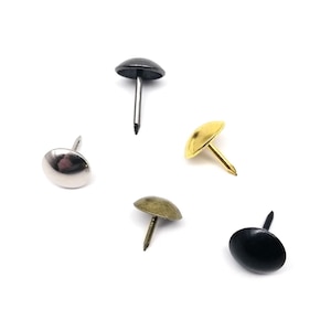 Puede incluir: Un surtido de tachuelas decorativas para tapicería en varios acabados metálicos, incluyendo plata, oro, bronce y negro. Cada tachuela presenta una cabeza redondeada y un alfiler afilado para insertar en tela u otros materiales. Las tachuelas están dispuestas sobre un fondo blanco.