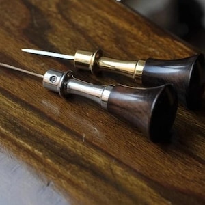 Op de afbeelding: Twee leerbewerkingspennen met donkerhouten handvatten en metalen accenten. Eén pen heeft een zilverkleurig metalen accent en de andere een goudkleurig metalen accent. De pennen liggen op een houten oppervlak.