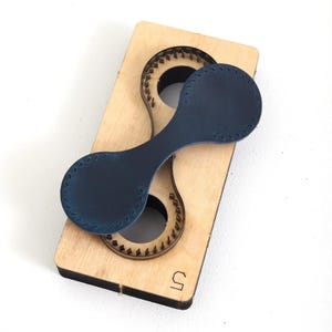 May include: A navy blue leather piece with stitched edges rests on a wooden die-cut form. The die-cut has a matching shape with a detailed edge. The wooden form has the number "5" etched on the bottom right corner. The background is white.