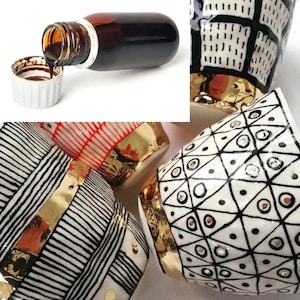 May include: A bottle of dark liquid is being poured into a white cap. Several ceramic mugs with black and white geometric patterns and gold accents are also visible. The mugs have unique designs, including stripes, triangles, and circles.