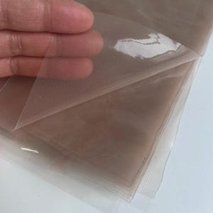 May include: A sheet of sheer, light beige mesh fabric with a fine, uniform hexagonal grid pattern. The material is translucent, lightweight, and flexible, showing a subtle texture across its surface when layered and held up to the light.