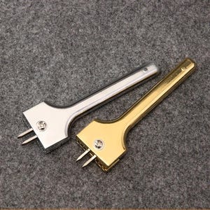 May include: Two metal leathercraft tools. One is silver-colored, the other is gold-colored. Each tool has a handle and a head with two prongs. The tools are designed for leatherworking and crafting.