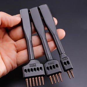 Puede incluir: Tres herramientas negras para artesanía en cuero con espaciado de 4 mm, sostenidas en una mano. Cada herramienta tiene una cabeza rectangular con múltiples púas de color cobre. Los mangos son largos y delgados, con un acabado mate.