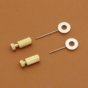 Könnte beinhalten: Zwei goldfarbene, strukturierte Schraubpfosten mit kleinem, geriffeltem Kopf und zwei silberfarbene, kreisförmige Stifte mit einem zentralen Loch. Die Artikel sind auf einer braunen Oberfläche angeordnet.
