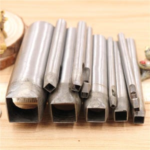 May include: A collection of eight silver metal leathercraft tools with square cutting ends. The tools are of varying sizes, with the largest on the left and the smallest on the right. The tools are arranged in a row, with the cutting ends facing forward.