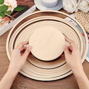 Puede incluir: Un conjunto de aros de bordado de madera y un disco redondo de madera. Los aros están anidados en varios tamaños, con un acabado de veta de madera clara. El disco está sostenido por manos y también está hecho de madera clara.