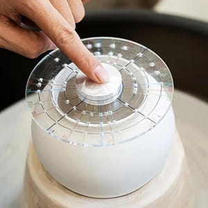 May include: A circular clear acrylic pottery centering tool with printed black grid lines and numerical markings, featuring a central silver-colored metal knob, positioned on top of a white clay vessel on a pottery wheel for precise alignment.