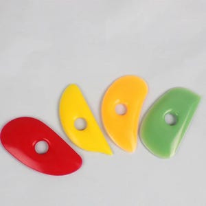 May include: A set of four kidney-shaped pottery ribs made of smooth, flexible plastic. The tools are coloured red, yellow, orange, and light green, each featuring a circular hole in the centre for a secure grip during clay shaping and smoothing tasks.