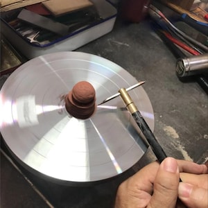 May include: A small, reddish-brown clay form centered on a circular, metallic spinning wheel. A metal tool with a brass-colored handle and a thin, pointed tip is positioned against the side of the clay to shape the rotating object.