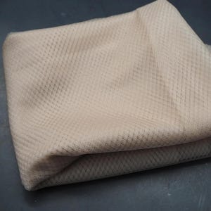 Pode incluir: Uma peça de tecido bege dobrada com uma construção de malha fina e semitransparente com um padrão de grade em forma de diamante consistente em todo o material, exibindo uma textura leve e respirável com variações tonais sutis na superfície.