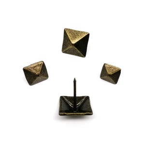 Puede incluir: Cuatro tachuelas y clavos decorativos de tapicería de color latón antiguo. Las tachuelas tienen una base cuadrada con un pasador central, mientras que los clavos tienen forma de pirámide. El metal tiene un acabado envejecido.