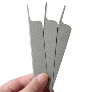 Puede incluir: Primer plano de tres cuchillas plateadas para cuchillo. Cada cuchilla tiene forma triangular con la parte superior curvada y una serie de líneas diagonales. Las cuchillas se mantienen juntas, sobre un fondo blanco.