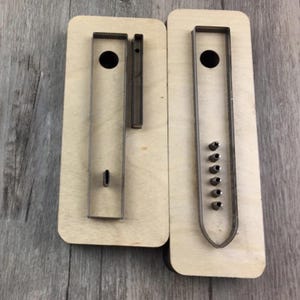 Könnte beinhalten: Zwei Lederhandwerkzeuge aus Holz mit Stanzformen aus Metall. Eines hat eine rechteckige Form mit einem kreisförmigen Loch und einem kleinen ovalen Loch. Das andere hat eine rechteckige Form mit einem kreisförmigen Loch und mehreren kleinen kreisförmigen Löchern.