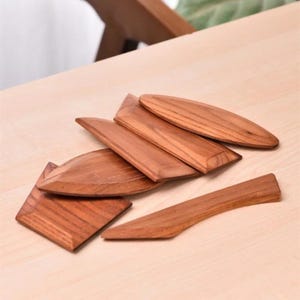 May include: A collection of six small, smooth, light brown wooden tools with visible grain patterns. The set includes various shapes such as a pointed oval, a rectangular piece with angled edges, and a long, slender, slightly curved handle-like tool.