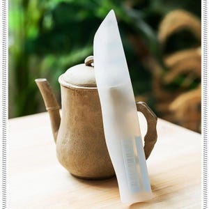 May include: A translucent white plastic knife sheath with a visible barcode and blue text markings, positioned vertically in front of a textured, light brown ceramic teapot with a lid and a curved handle.