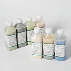 Puede incluir: Siete botellas de plástico transparente llenas de diversos materiales de barbotina o esmalte cerámico. Cada botella cuenta con una tapa de rosca blanca y una etiqueta rectangular blanca con texto negro y un borde verde informativo.