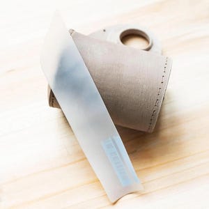 May include: A cylindrical beige leather sleeve with visible stitched edges and a circular cutout at the top, partially covered by a translucent white plastic strip featuring a small blue barcode label near the bottom edge of the strip.