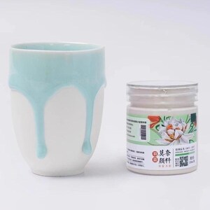 Puede incluir: Una taza de cerámica blanca con un esmalte azul claro que gotea desde el borde, junto a un frasco cilíndrico de plástico de pigmento cerámico con una etiqueta que muestra una flor blanca y texto que incluye 100ml.