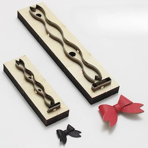 Può includere: Strumenti di taglio in legno per creare fiocchi. Due blocchi di legno con ritagli metallici a forma di fiocco, insieme a fiocchi in pelle rossa e nera finiti. Ideale per lavori in pelle e progetti fai-da-te.