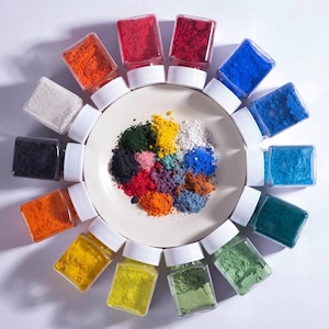 May include: A circular white plate sits in the center surrounded by fourteen clear square plastic jars with white lids, each filled with a different colored pigment powder. The plate contains small piles of various colored powders in a circular arrangement.
