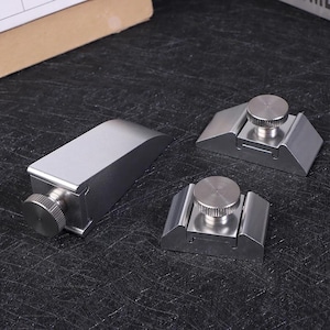 Puede incluir: Tres prensas de carpintería de metal plateado con perillas ajustables. Una prensa es rectangular con la parte superior curvada, y dos son más pequeñas, con base plana y perilla circular. Sobre una superficie oscura.