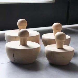 Puede incluir: Cuatro herramientas de madera de color claro con bases redondeadas en forma de cuenco y mangos verticales centrales. Cada pieza presenta una superficie lisa y sin acabado con patrones de veta de madera natural, dispuestas en un grupo variado.