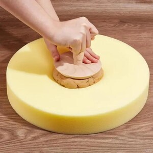 Op de afbeelding: Een persoon gebruikt een houten gereedschap om klei te vormen op een gele schuimbasis. Het gereedschap heeft een ronde houten handgreep en een cirkelvormige basis. De klei is lichtbruin van kleur. De schuimbasis is cirkelvormig.