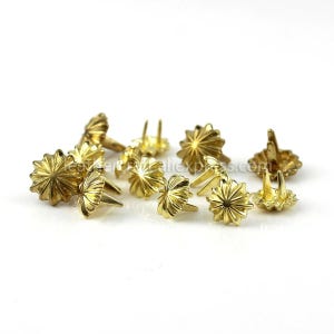 May include: A collection of gold-colored upholstery tacks. Each tack features a decorative, starburst-shaped head and three prongs for secure attachment. These tacks are likely used for furniture or decorative projects.