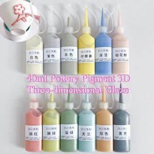 Puede incluir: Doce botellas exprimibles de 40 ml de esmalte de pigmento para cerámica 3D en varios colores, cada una con una punta aplicadora puntiaguda y una etiqueta con caracteres chinos. Las botellas están dispuestas en dos filas, mostrando blanco, morado, gris, amarillo, azul, rosa, rojo, verde claro, verde oscuro, azul claro, marrón y negro.